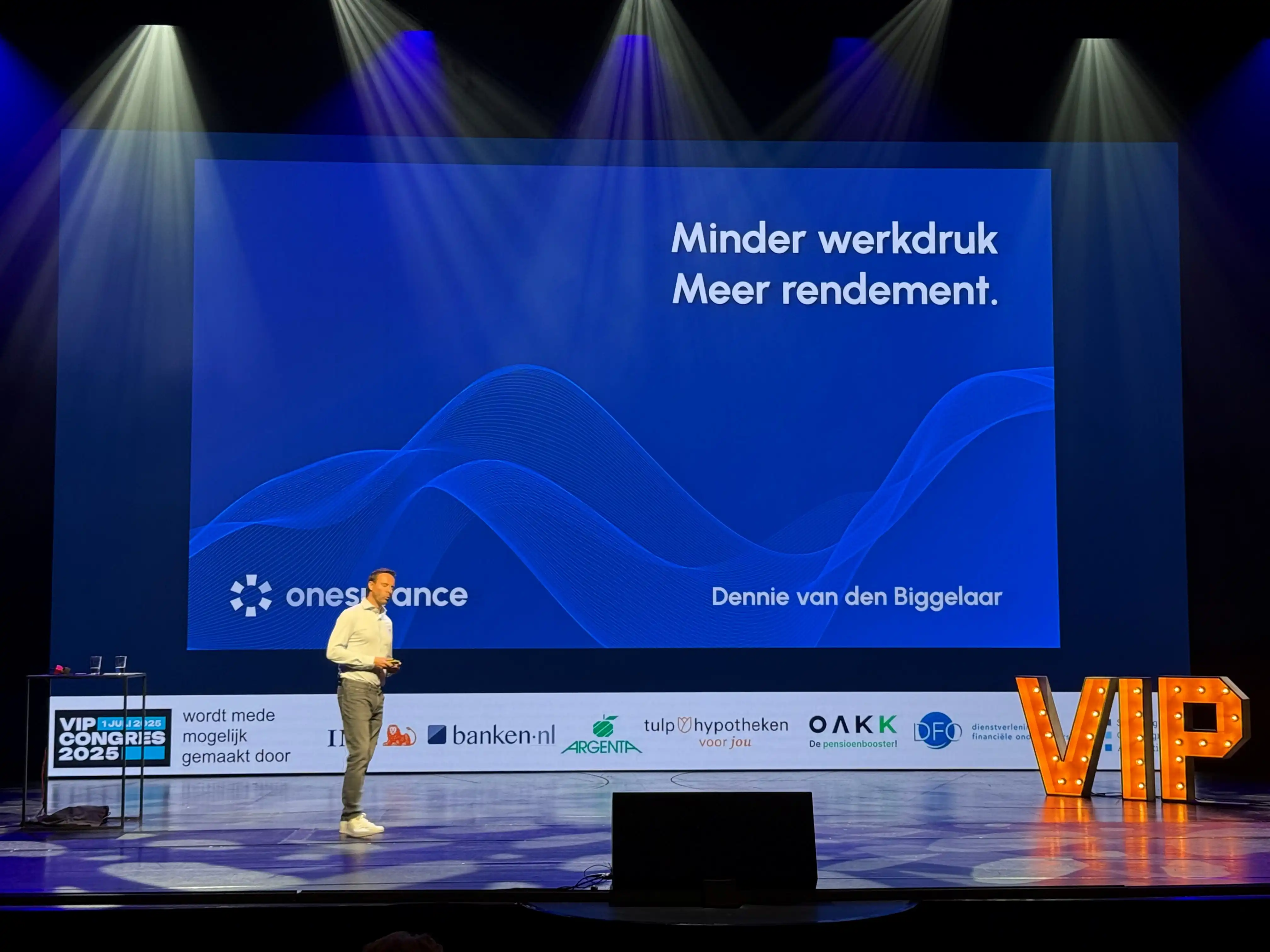 Dennie van den Biggelaar op het podium van het VIP Congres 2025 in het AFAS Theater Leusden, met op het scherm: Minder werkdruk, meer rendement.